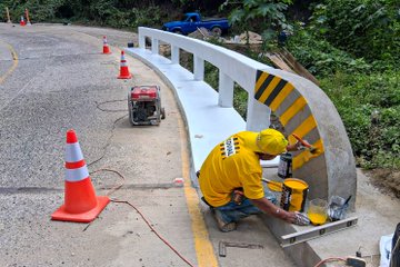 Multiple images show construction workers in yellow vests and helmets performing maintenance on a concrete bridge railing along a roadside surrounded by green vegetation and orange traffic cones. One image depicts workers applying plaster or cement to curved railing sections using trowels and buckets of material, with tools like brooms and a generator nearby. Another focuses on a completed curved concrete railing base adjacent to lush greenery and a road. A third image captures a worker painting yellow and black stripes on the railing with a brush and paint can, positioned next to a blue truck and more cones. The final image illustrates a finished white bridge with yellow safety markings over a small stream, featuring a motorcyclist crossing it amid surrounding trees and cones.