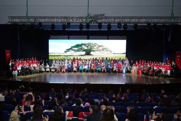 First image shows a large group of adults and children standing on a stage in formal attire including suits, dresses, and red clothing, with Turkish flags and banners in the background, a wooden table holding flowers in the foreground. Second image depicts rows of schoolchildren in colorful traditional and modern outfits standing on a stage with a large screen displaying a tree in a field, Turkish flags on sides, audience seated below. Third image features ministers and officials in suits and red attire posing with smiling children wearing red shirts and Turkish flag motifs, cameras and audience in background. Fourth image captures a group of people in various outfits including red scarves and suits on a stage with banners reading about Republic Day event, Turkish flags and microphones present.