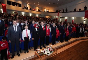 First image shows a large group of adults and children standing on a stage in formal attire including suits, dresses, and red clothing, with Turkish flags and banners in the background, a wooden table holding flowers in the foreground. Second image depicts rows of schoolchildren in colorful traditional and modern outfits standing on a stage with a large screen displaying a tree in a field, Turkish flags on sides, audience seated below. Third image features ministers and officials in suits and red attire posing with smiling children wearing red shirts and Turkish flag motifs, cameras and audience in background. Fourth image captures a group of people in various outfits including red scarves and suits on a stage with banners reading about Republic Day event, Turkish flags and microphones present.