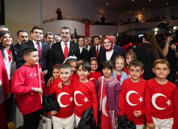 First image shows a large group of adults and children standing on a stage in formal attire including suits, dresses, and red clothing, with Turkish flags and banners in the background, a wooden table holding flowers in the foreground. Second image depicts rows of schoolchildren in colorful traditional and modern outfits standing on a stage with a large screen displaying a tree in a field, Turkish flags on sides, audience seated below. Third image features ministers and officials in suits and red attire posing with smiling children wearing red shirts and Turkish flag motifs, cameras and audience in background. Fourth image captures a group of people in various outfits including red scarves and suits on a stage with banners reading about Republic Day event, Turkish flags and microphones present.