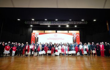 First image shows a large group of adults and children standing on a stage in formal attire including suits, dresses, and red clothing, with Turkish flags and banners in the background, a wooden table holding flowers in the foreground. Second image depicts rows of schoolchildren in colorful traditional and modern outfits standing on a stage with a large screen displaying a tree in a field, Turkish flags on sides, audience seated below. Third image features ministers and officials in suits and red attire posing with smiling children wearing red shirts and Turkish flag motifs, cameras and audience in background. Fourth image captures a group of people in various outfits including red scarves and suits on a stage with banners reading about Republic Day event, Turkish flags and microphones present.