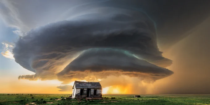 A huge storm cloud swirls above a wide open plain, with a small broken-down shack in the foreground.