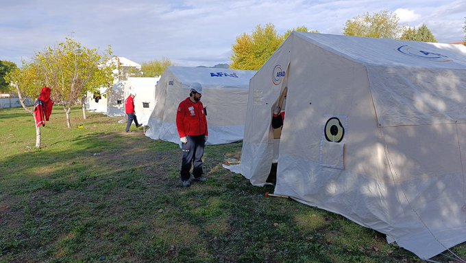 First image shows several white tents with MEB AKUB logos set up on a grassy field surrounded by autumn trees with yellow leaves under a partly cloudy sky. Personnel in red jackets and helmets stand near the tents some entering or exiting one with a red flag nearby. Second image depicts a large group of about 20-25 people mostly in red uniforms and helmets kneeling or standing in front of similar white tents on the grass with trees in the background.