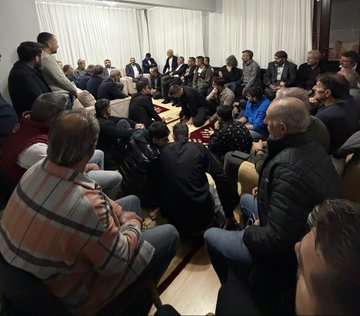First image shows a group of about 20 men of various ages sitting in a circle on the floor around a red rug in a room with white curtains and wooden floor, some wearing jackets and others casual clothes, engaged in discussion. Second image depicts a similar indoor gathering of men seated on the floor and chairs around a red rug, with one man standing and speaking, room has white walls and curtains. Third image features men sitting on beige sofas and floor around a red rug in a lit room with white curtains and a TV on the wall, some in suits and others in casual attire. Fourth image displays a large group of over 30 men standing in a semi-circle outdoors at night near trees and a black car, illuminated by streetlights, wearing coats and jackets, posing for a group photo.