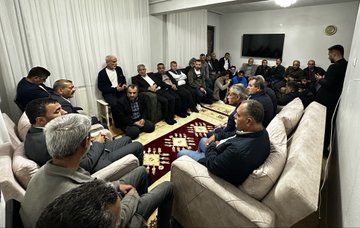 First image shows a group of about 20 men of various ages sitting in a circle on the floor around a red rug in a room with white curtains and wooden floor, some wearing jackets and others casual clothes, engaged in discussion. Second image depicts a similar indoor gathering of men seated on the floor and chairs around a red rug, with one man standing and speaking, room has white walls and curtains. Third image features men sitting on beige sofas and floor around a red rug in a lit room with white curtains and a TV on the wall, some in suits and others in casual attire. Fourth image displays a large group of over 30 men standing in a semi-circle outdoors at night near trees and a black car, illuminated by streetlights, wearing coats and jackets, posing for a group photo.