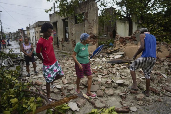 Group of people including a young Black man in red shirt and shorts, an older woman in green top and headscarf holding an umbrella, and an older man in blue tank top and checkered shorts stand on a debris-strewn dirt road amid ruined colonial-style buildings with collapsed walls and exposed interiors, surrounded by uprooted trees, scattered rubble, power lines, and a rainy overcast sky in a Caribbean street setting.
