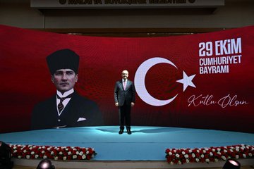 First image shows a large group of formally dressed men and women including officials in suits and military uniforms some wearing headscarves gathered around a round table covered with white cloth set with glasses bottles of beverages plates of food and flower arrangements in a grand hall decorated with red Turkish flags hanging from the ceiling and walls. Second image depicts a stage with a large red backdrop displaying a portrait of Mustafa Kemal Atatürk in traditional attire next to a modern suited man with text reading 29 EKİM CUMHURİYET BAYRAMI and KUTLU OLSUN surrounded by blue flooring and red carpet with white flowers on either side. Third image features six men in suits and uniforms standing on a stage in front of a similar red banner with Atatürk portrait and Republic Day text blue floor and flower arrangements emphasizing formal poses. Fourth image captures an older man in a dark suit with red tie kneeling beside a younger man in a wheelchair wearing a light jacket both smiling and holding hands in a warmly decorated room with flags balloons tables and other attendees in background.