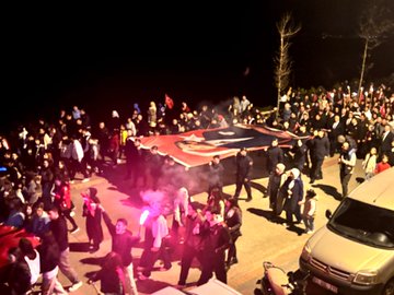 First image shows overhead view of nighttime crowd with multiple large Turkish flags waving trees and lights in background. Second image captures group carrying red flag with white crescent and star smoke and purple lights visible van nearby. Third image depicts line of men in suits holding Turkish flags building and banners behind them. Fourth image features long red banner with white emblem held by walking participants trees vehicles and green lights along street.