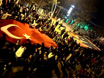 First image shows overhead view of nighttime crowd with multiple large Turkish flags waving trees and lights in background. Second image captures group carrying red flag with white crescent and star smoke and purple lights visible van nearby. Third image depicts line of men in suits holding Turkish flags building and banners behind them. Fourth image features long red banner with white emblem held by walking participants trees vehicles and green lights along street.
