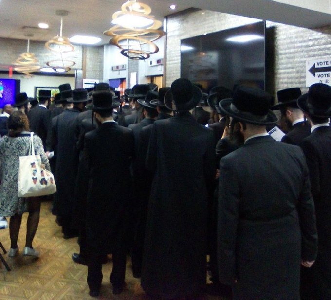 A group of men wearing black hats and suits stands in a line inside a room with beige brick walls and multiple hanging pendant lights. A large flat screen television is mounted on the wall displaying a vote sign. A woman in a patterned dress carrying a white tote bag stands nearby. The floor is wooden with a chair and small table in the foreground.