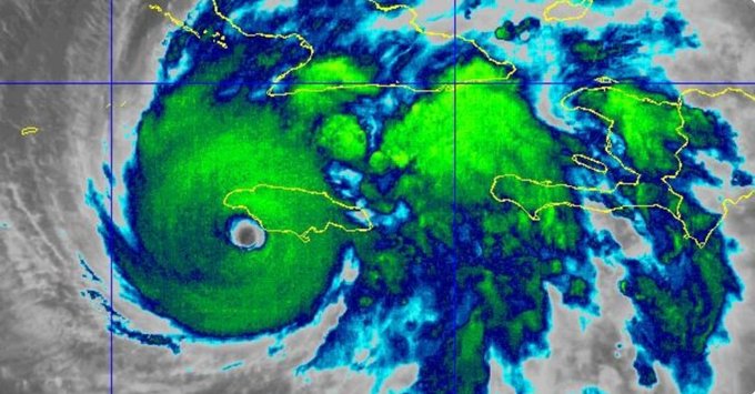 Satelite image displays a large swirling hurricane system over the Atlantic Ocean near the Caribbean islands, with green and yellow colors indicating intense storm clouds and rain bands, a clear eye at the center, and outlined landmasses including parts of Cuba, Jamaica, Haiti, and the Dominican Republic visible in the surrounding areas.