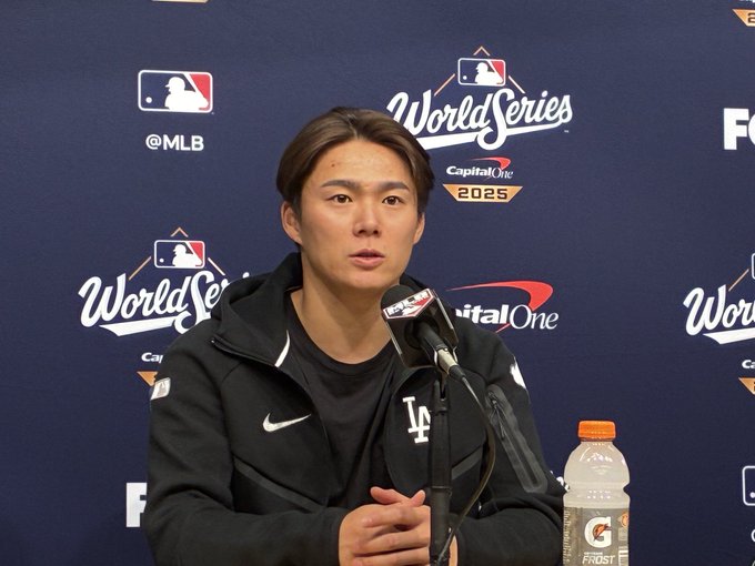 A Japanese man with short black hair wearing a black zip-up jacket with Dodgers logo and black shirt stands at a podium holding a microphone during a press conference. He has a focused expression while speaking. Behind him is a blue backdrop featuring World Series and MLB logos along with Capital One sponsorship text and 2025 year marking. A bottle of Gatorade is placed on the podium to his right.