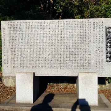 First image shows panoramic landscape with blue sky over distant sea city buildings hills and green forested areas in foreground. Second image depicts tall abstract bronze statue resembling flames on stone pedestal amid trees shrubs and paved area. Third image features large rectangular gray stone monument engraved with dense Japanese text characters on front and sides supported by two bases with shadows. Fourth image displays wide stone staircase ascending between green bushes pine trees and stone lanterns leading to elevated paved platform under clear sky.