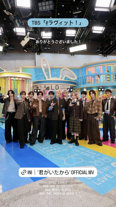 Eleven young men identified as INI group members stand in a group pose on a vibrant TV studio set with blue bunny ear structures yellow floor stripes and colorful backgrounds. They wear matching school uniforms including brown blazers white shirts plaid skirts for one member and trousers in beige and black. Bright studio lights illuminate the scene and overlaid text reads TBS logo Japanese for Ravit show and INI Rocketeer Official MV promotion.