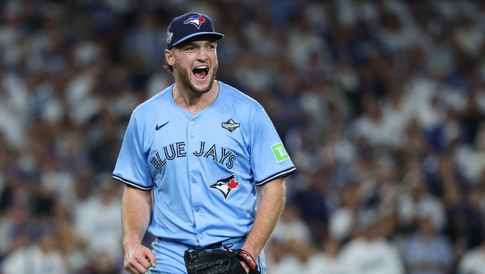 A male baseball player wearing a blue Toronto Blue Jays uniform jersey with Nike logo, TLN sponsor patch, and team emblem on the sleeve, along with a matching blue cap, stands on a baseball field during a game, smiling widely with mouth open, holding a baseball glove on his right hand, surrounded by a blurred crowd of spectators in the stadium stands.