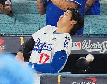Shohei Ohtani wears a white Los Angeles Dodgers jersey with number 17, blue accents, Nike swoosh, MLB logo, and World Series patch, leaning back with arms extended to avoid an incoming white baseball near protective netting in a stadium dugout area. Spectators in blue and white attire watch from seats behind the net, with a blurred person in a blue cap nearby. Blue Dodgers logo and Capital One advertisement appear on the yellow railing below.