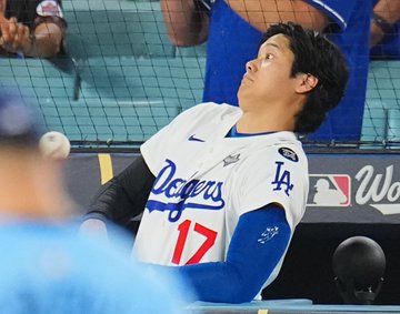 Shohei Ohtani wears a white Los Angeles Dodgers jersey with number 17, blue accents, Nike swoosh, MLB logo, and World Series patch, leaning back with arms extended to avoid an incoming white baseball near protective netting in a stadium dugout area. Spectators in blue and white attire watch from seats behind the net, with a blurred person in a blue cap nearby. Blue Dodgers logo and Capital One advertisement appear on the yellow railing below.