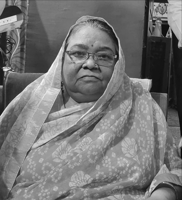An elderly Indian woman with a serious expression sits on a cushioned chair in a simple indoor setting with a window and lamp nearby. She wears a white saree with floral patterns draped as a dupatta over her head, round glasses, a nose ring, earrings, and a necklace. Her hair is gray and tied back.