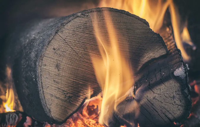 Close-up view of a large log of wood with rough bark partially burned by orange and yellow flames licking its surface in a dark indoor setting suggesting a fireplace the flames illuminate the wood grain and emit sparks
