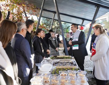 First image shows a man in a suit and keffiyeh smiling and hugging a group of young students in green shirts with school logos, standing in front of a banner reading Bir Okul Bin Umut Gazze, under trees in an outdoor school setting. Second image depicts the same man in suit and keffiyeh interacting with a diverse group of adults and youth near a food stand with trays of pastries, cookies, and dishes on a table, outdoors with autumn foliage and a metal shelter. Third image features the man in suit and keffiyeh standing with students in green shirts beside a trophy on a table inside a building with Turkish flag, a red box, and award plaques visible. Fourth image displays a large group of people in white lab coats and suits posing in front of the Yesil Teknoloji Mesleki ve Teknik Anadolu Lisesi building entrance on a sunny day.