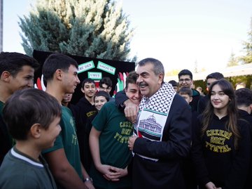 First image shows a man in a suit and keffiyeh smiling and hugging a group of young students in green shirts with school logos, standing in front of a banner reading Bir Okul Bin Umut Gazze, under trees in an outdoor school setting. Second image depicts the same man in suit and keffiyeh interacting with a diverse group of adults and youth near a food stand with trays of pastries, cookies, and dishes on a table, outdoors with autumn foliage and a metal shelter. Third image features the man in suit and keffiyeh standing with students in green shirts beside a trophy on a table inside a building with Turkish flag, a red box, and award plaques visible. Fourth image displays a large group of people in white lab coats and suits posing in front of the Yesil Teknoloji Mesleki ve Teknik Anadolu Lisesi building entrance on a sunny day.