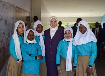 First image shows a group of young girls and women wearing white headscarves and blue school uniforms standing together in a hallway with some men in suits nearby. Second image depicts a classroom scene with teachers including women in headscarves and a man in a suit standing at the front near a projector screen while students in blue uniforms sit at desks. Third image features a large group of adults in formal attire and young children in colorful uniforms sitting in a circle on the floor in a classroom decorated with sun and animal drawings. Fourth image displays a diverse group of children and adults in traditional African clothing and headwraps posing outdoors under trees with decorative hats and water bottles visible.