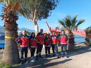 Group of six rescuers in red uniforms and helmets stand together on sandy shore near palm trees and a red bridge over water with a rescue boat visible in background. Wide view shows red bridge over lake with boats rescuers in orange suits entering water and crowd on shore during drill. Three rescuers in red gear handle equipment like hoses near red bridge over water with mountains in distance. Two rescuers in red uniforms observe from shore as others in boats and water conduct operations near blue tarp during exercise.