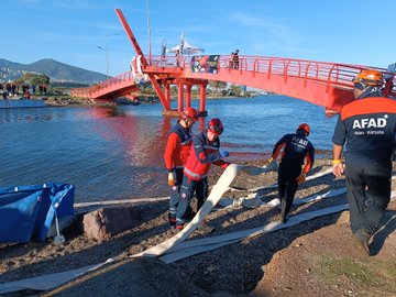 Group of six rescuers in red uniforms and helmets stand together on sandy shore near palm trees and a red bridge over water with a rescue boat visible in background. Wide view shows red bridge over lake with boats rescuers in orange suits entering water and crowd on shore during drill. Three rescuers in red gear handle equipment like hoses near red bridge over water with mountains in distance. Two rescuers in red uniforms observe from shore as others in boats and water conduct operations near blue tarp during exercise.