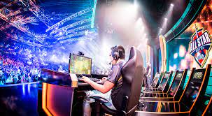 A gamer wearing a headset sits at a desk with multiple monitors and keyboards in a brightly lit arena stage setup featuring large screens and the All-Star logo banners surrounded by other similar gaming stations and audience seating areas.