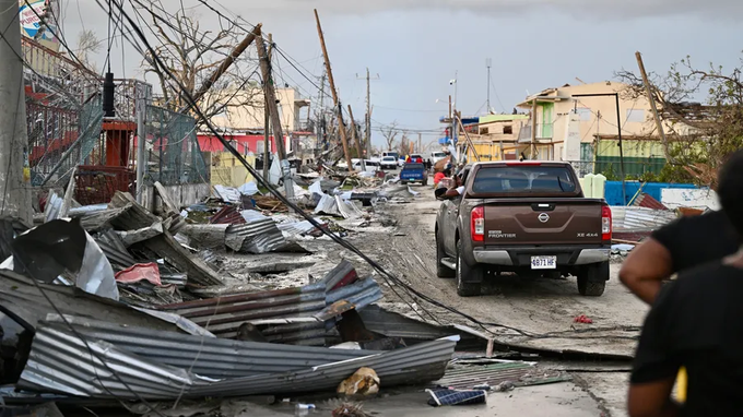 Street scene in a damaged urban area with scattered metal debris fallen utility poles and wires wrecked buildings and fences a brown Nissan truck parked amid rubble people in dark clothing observing the destruction under overcast skies with broken trees and scattered trash visible