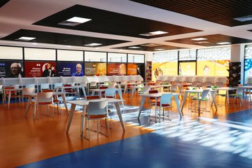 First image shows a group of men and women in formal business attire, including suits and dresses, seated in rows on chairs facing forward, some wearing name badges, in an indoor setting with a microphone stand visible. Second image depicts a larger group of men and women in suits and dresses standing in a semi-circle on an orange-tiled floor under a ceiling with lights, in front of a white table and chair. Third image displays an empty modern library interior with orange and blue patterned flooring, white round tables and chairs arranged in rows, large windows with colorful wall displays and banners featuring text and images.