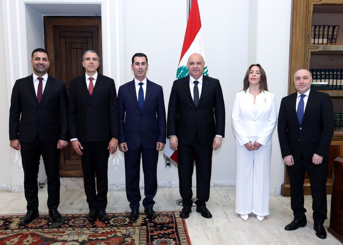 Six people stand in a formal group pose inside an ornate room with wooden doors and a large Lebanese flag on the wall. Five men wear dark suits with ties in various colors and the woman wears a white dress and jacket. They position themselves in two rows with hands at sides or clasped, on a patterned rug with bookshelves visible in the background.