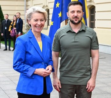 First image shows a man with short gray hair wearing a dark suit and blue tie, speaking into a microphone while gesturing with his hand, standing in front of a blue backdrop with the flags of Slovakia and Russia visible. Second image depicts three people standing outdoors near a building: a woman with short gray hair in a blue blazer and yellow top smiling, a man with dark hair in a green military-style polo shirt, and European Union flags in the background.