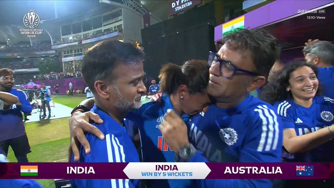 Image captures Captain Harmanpreet Kaur in blue India team jersey hugging a female teammate also in blue jersey on green cricket field after victory, both showing emotional expressions with tears, surrounded by other players in blue jerseys celebrating, stadium seating and purple lighting in background, scoreboard displays India won by wickets against Australia with flags.