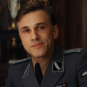 A black-and-white historical photograph of a man in a Nazi SS uniform, featuring a high-ranking officer with a serious expression, wearing a peaked cap, epaulettes, and insignia including swastika armbands and Iron Cross medals; the background shows ornate wooden paneling and decorative vases, suggesting an indoor formal setting.