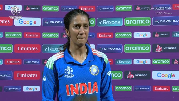 A woman in an Indian cricket team jersey stands at a press conference podium with the word INDIA prominently displayed on her uniform. She has long dark hair and wears a focused expression. The backdrop features numerous sponsor logos including Google, DP World, Aramco, Emirates, Rexona, and Xface in various colors against a purple background. The setting appears to be a professional sports event with branding for the ICC Womens Cricket World Cup.