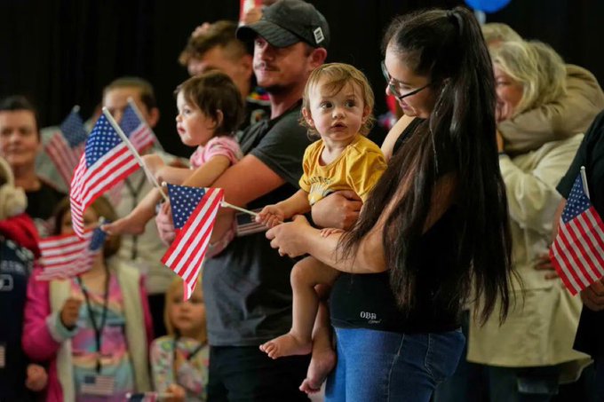 A crowded indoor event with a diverse group of people, many holding small American flags, including families with young children in yellow outfits; attendees appear to be at a patriotic rally or gathering, with some wearing casual clothing and others bundled up, suggesting a public assembly like a Trump supporter event.