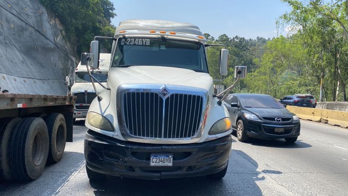 White semi-truck trailer with license plate C-234BYB stopped on a multi-lane highway uphill section near a rocky cliffside and green trees. Black Mazda sedan visible in adjacent lane. Additional trucks and barriers in background under clear sky.