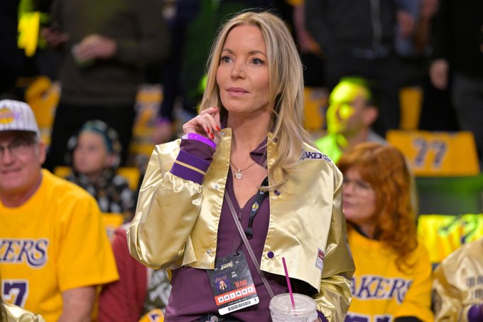 A blonde woman with long hair sits in a basketball stadium, wearing a shiny gold jacket with purple cuffs and Lakers branding, a black top underneath, a lanyard with a badge around her neck, and holding a clear plastic cup with a straw. She has a thoughtful expression, finger on her chin. Surrounding her are spectators including a man in glasses wearing a yellow Lakers shirt, a red-haired woman in a purple top, and others in yellow shirts and hats, with seat numbers like 77 visible and a green glow in the background.