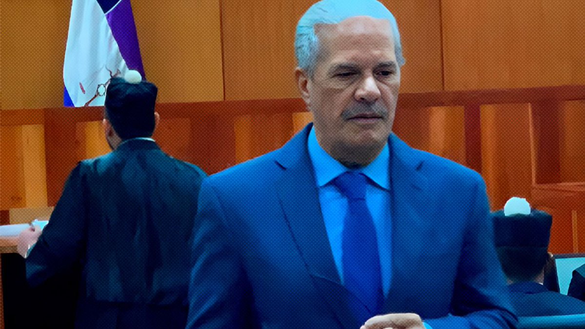 Two men in dark suits stand in a wooden-paneled room with a Dominican Republic flag on the wall one man faces forward wearing a blue suit and blue tie with white hair the other man stands behind with head turned partially visible desks and chairs in background