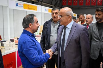 First image shows a man in a blue shirt shaking hands with a man in a gray suit and tie in front of book stacks and fair booths with Turkish flags and banners including Malatya Ticaret Borsası. Second image depicts a group of men in suits and casual attire gathered around discussing near book displays and exhibition stands with Turkish flags in a large indoor fair hall. Third image features men in formal wear including suits and vests shaking hands and standing near book stalls with promotional posters and Turkish flags at the event. Fourth image displays several men in suits standing together in front of stacked red and white books and fair booths labeled with numbers and Malatya Ticaret Borsası in a spacious exhibition area.