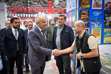 First image shows a man in a blue shirt shaking hands with a man in a gray suit and tie in front of book stacks and fair booths with Turkish flags and banners including Malatya Ticaret Borsası. Second image depicts a group of men in suits and casual attire gathered around discussing near book displays and exhibition stands with Turkish flags in a large indoor fair hall. Third image features men in formal wear including suits and vests shaking hands and standing near book stalls with promotional posters and Turkish flags at the event. Fourth image displays several men in suits standing together in front of stacked red and white books and fair booths labeled with numbers and Malatya Ticaret Borsası in a spacious exhibition area.