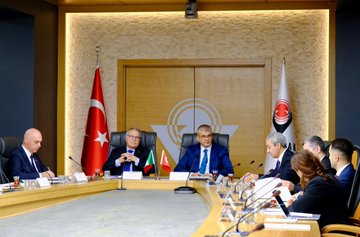 First image shows two men in suits standing at podiums holding blue and red folders with logos one with glasses wearing a blue tie and the other in a blue suit with a blue tie Turkish and Italian flags on either side wooden doors in background. Second image depicts a long conference table with multiple suited men and women seated around it including bald man and woman with glasses Turkish and Italian flags and TÜBİTAK logo on walls modern room with lighting. Third image features a group of eight suited men and women standing in front of large screens displaying Turkish Italian and another flag wooden table with microphones below. Fourth image shows five people seated around an oval table in suits including bald man and woman with headscarf Turkish Italian and other flags in background conference setup with documents.