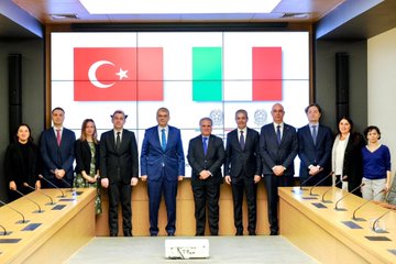 First image shows two men in suits standing at podiums holding blue and red folders with logos one with glasses wearing a blue tie and the other in a blue suit with a blue tie Turkish and Italian flags on either side wooden doors in background. Second image depicts a long conference table with multiple suited men and women seated around it including bald man and woman with glasses Turkish and Italian flags and TÜBİTAK logo on walls modern room with lighting. Third image features a group of eight suited men and women standing in front of large screens displaying Turkish Italian and another flag wooden table with microphones below. Fourth image shows five people seated around an oval table in suits including bald man and woman with headscarf Turkish Italian and other flags in background conference setup with documents.