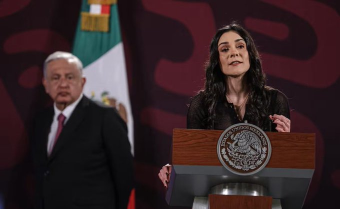 An older man in a dark suit with red tie stands to the left, partially visible, next to a woman with long wavy dark hair wearing a black top, speaking at a wooden podium bearing the Mexican presidential seal. Mexican flags are positioned behind them against a red and green backdrop with gold accents. The setting appears to be an official press conference room.
