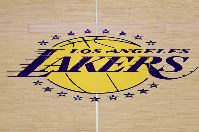 A wooden basketball court floor with white lines marking the center circle and half-court line. The purple and yellow Los Angeles Lakers logo is prominently displayed in the center, featuring a basketball with stars around it and the team name in bold letters.