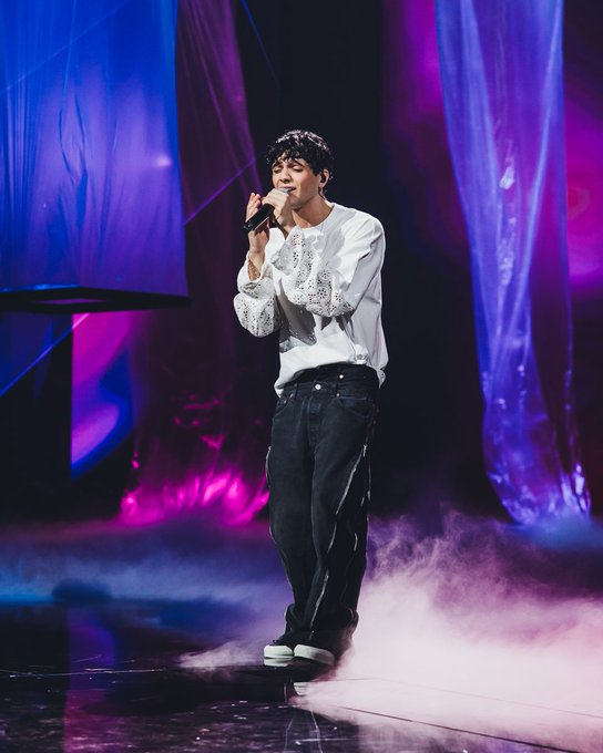 Young male performer with curly black hair stands on stage holding microphone close to mouth while singing. He wears white long-sleeve shirt with lace details on cuffs tucked into wide-leg black jeans and white sneakers. Blue and purple spotlights illuminate the scene with draped purple fabric backdrops and ground-level fog creating a misty effect around his feet. The setting appears to be a live music performance area.