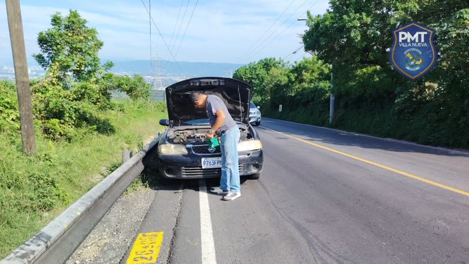 A dark sedan with its hood open is parked on the edge of a paved road in a rural setting with green grass, trees, and distant mountains under a clear sky. A man wearing a gray shirt, blue pants, and gloves stands beside the vehicle, appearing to inspect or repair the engine. A police patch is visible on his clothing, and another vehicle is partially seen in the background. Yellow road markings and a utility pole are present nearby.