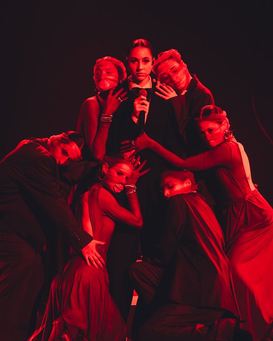 Group of performers in dark elegant clothing with red lighting central female figure in black outfit holding microphone surrounded by figures with face masks and expressive poses some wearing bangles and dresses arranged in clustered formation on dark background