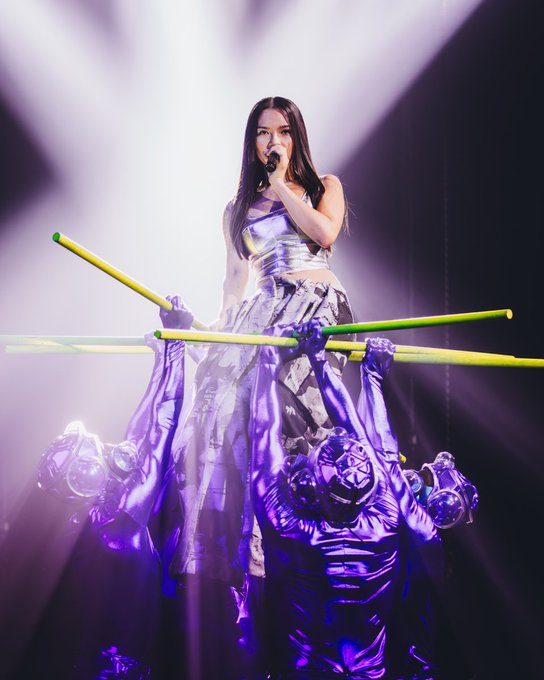 Singer Mayu stands center stage in a metallic silver sleeveless top and skirt holding yellow drumsticks with both hands. She has long dark hair and is singing into a microphone. Multiple dancers in purple outfits and face paint lift and surround her from below creating a dynamic group formation. Bright white spotlights illuminate the scene with a dark background.